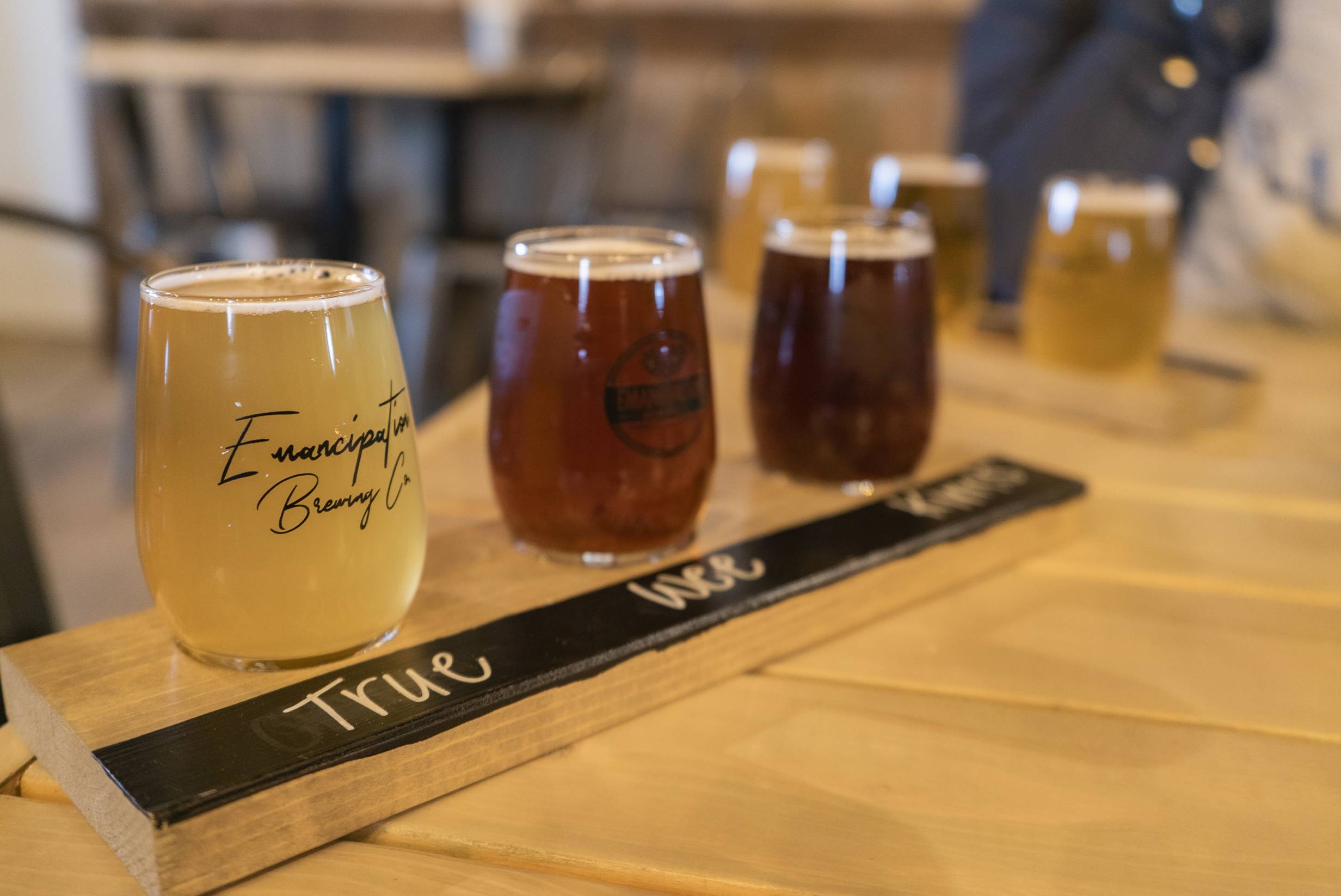 A flight of three beers at Emancipation Brewing. The first beer is light yellow, the second is light brown, and the third is a darker, reddish brown. The serving tray reads 