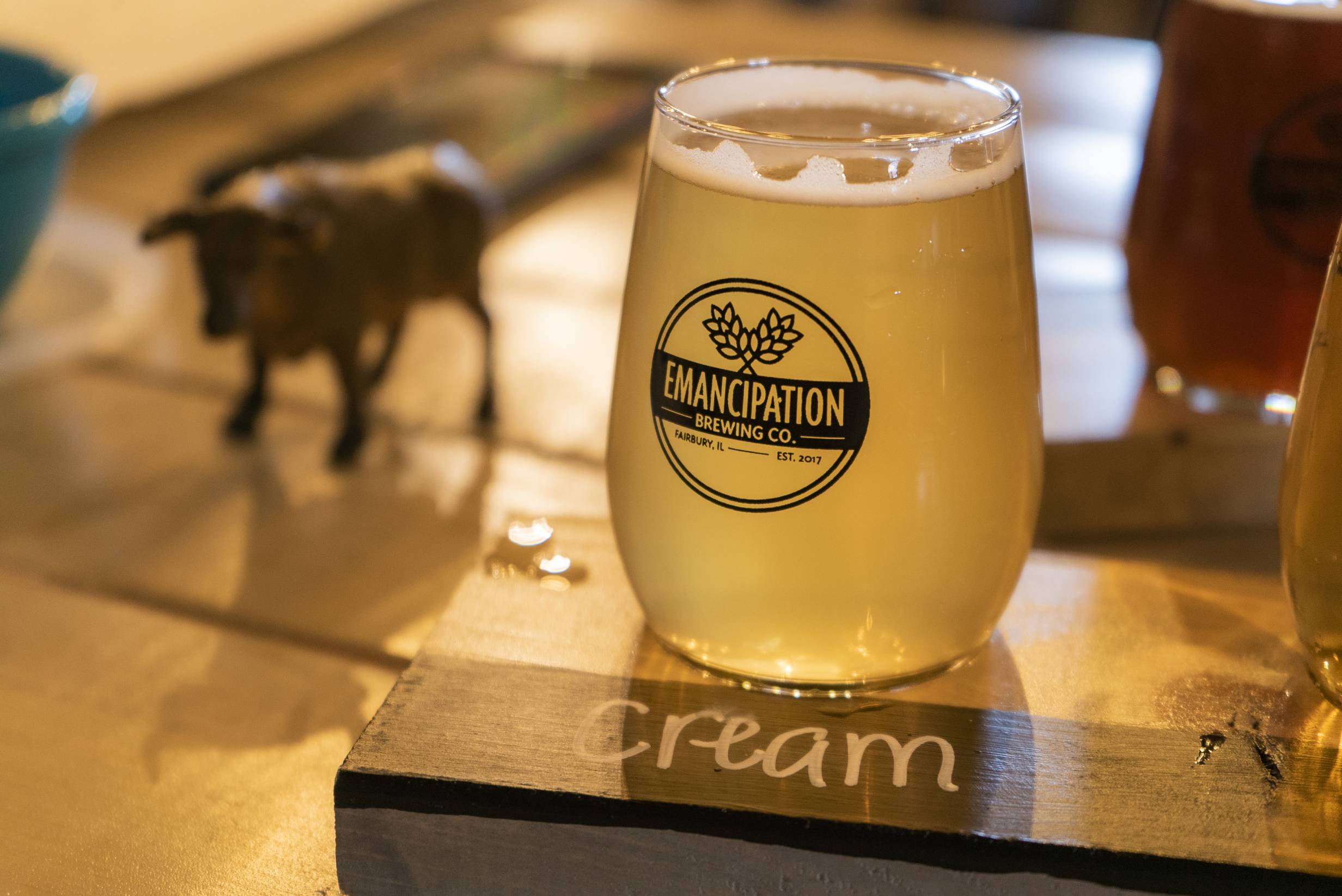 At Emancipation Brewing, a stemless tumbler glass with the brewery's logo on the front is filled with a light yellow beer. It sits on a wood serving tray, and the tray reads 