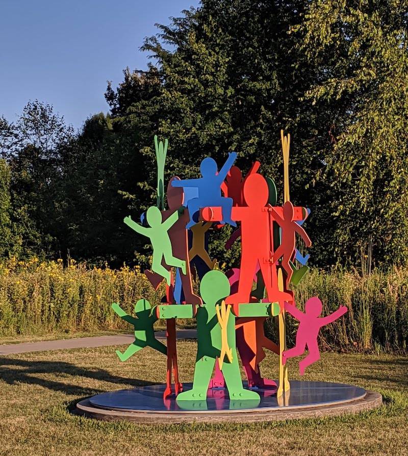 A metal structure of different colored simple human figures. There are larger figures creating a tower in the center, and smaller figures hanging off of that tower. The structure is surrounded by prairie grasses and trees. Photo by Katriena Knights.