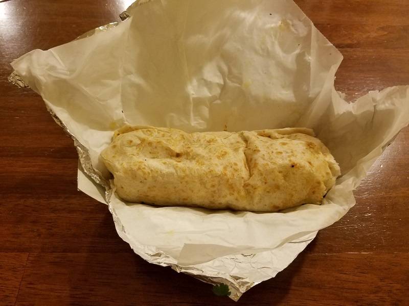 Burrito from Fernando's food truck. Only the white flour tortilla is visible. The burrito sits in unwrapped wax and tin foil on a reddish wood table. Photo by Matthew Macomber. 