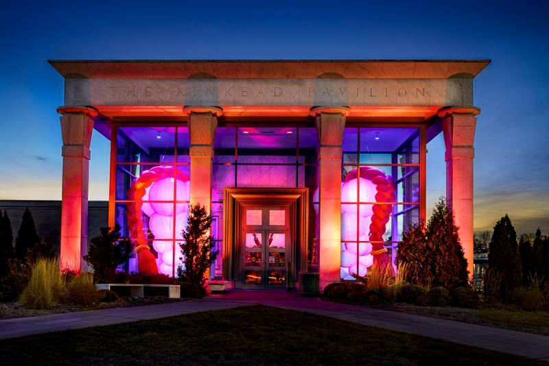 A view of the front entrance of Krannert Art Museum. The facade is mostly glass, and there are four columns in front. It is lit in orange and purple, and there are two red inflatable pieces visible through the glass. Photo by Fred Zwicky from Illinois News Bureau.