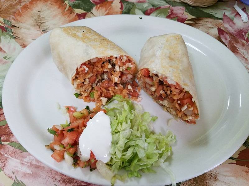 Burrito at Huaraches Moroleon is sliced in half. The inside of the burrito includes rice, tomatoes, chicken, and beans. It is on an oval white plate. On the plate is a small sald consisting of pico de gallo, sour cream, and shredded lettuce. Photo by Matthew Macomber. 