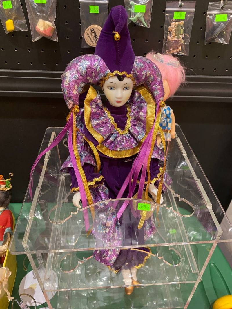 Image: A jester doll sits in a clear container. It has a costume in shades of purple and gold, with a ribbons coming out of two of the points of the jester hat. Photo by Julie McClure.