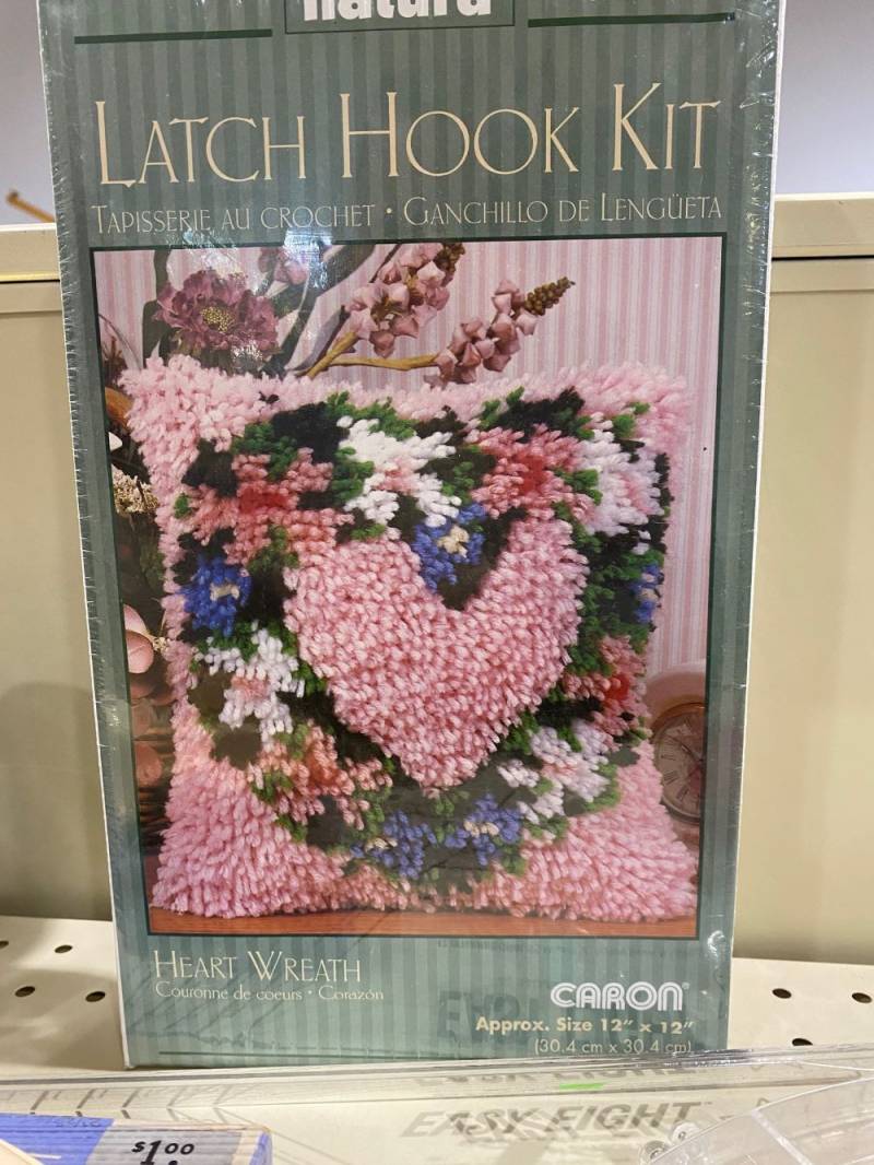 Image: A box sits on a metal shelf. It has green stripes and a photo of a pillow that has been latch hooked with a flowery heart and pink background. It says 