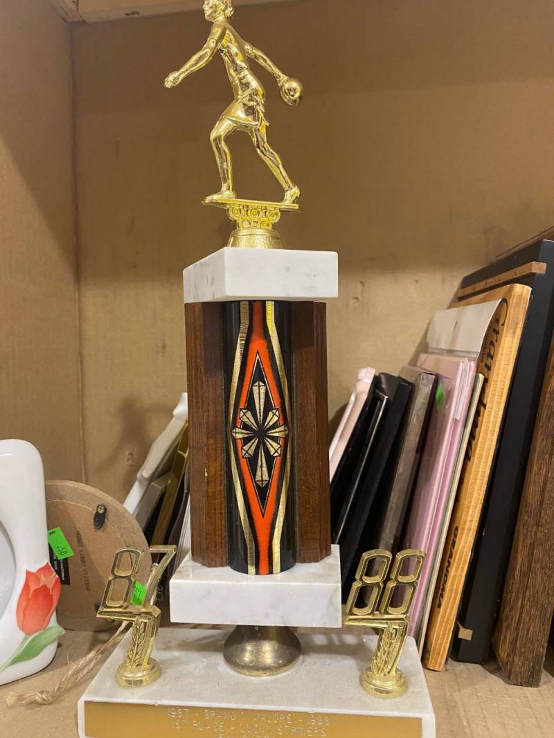 Image: A trophy sits on a shelf. It has a white base with a gold metal plate on the front. A woman bowler is at the top of the trophy, and the main part of the trophy is flanked by decorations that say 