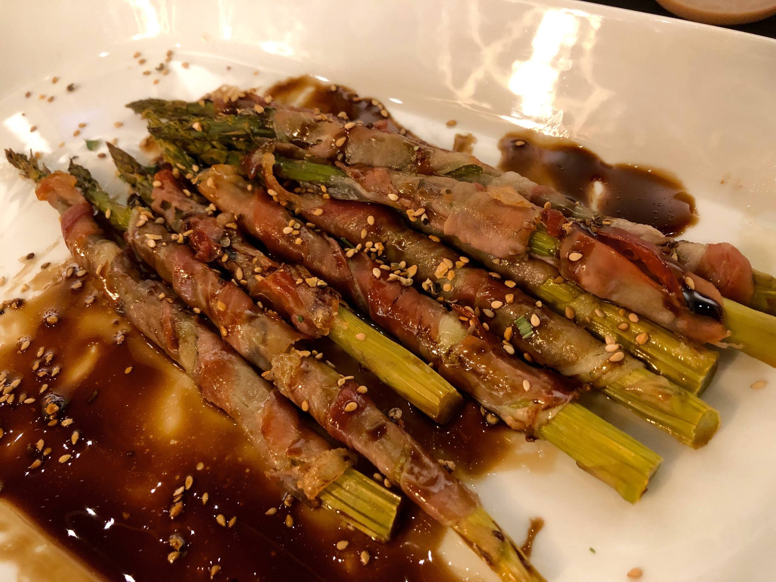 Image: Proscuitto-wrapped asparagus drizzled with balsamic vinegar and garnished with sesame seeds are served on white plate. The asparagus are a light green, and the vinegar is a thick, dark brown. Photo by Rebecca Wells. 