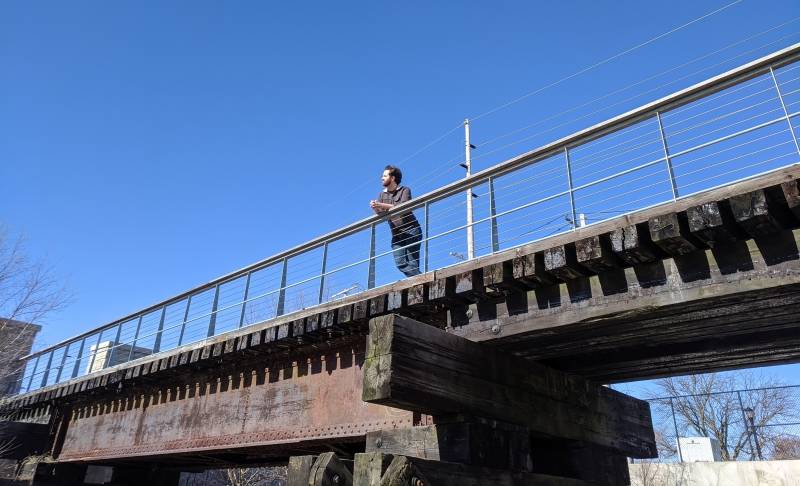 Image: The writer is standing on a bridge with a metal railing, with a blue sky backdrop. Photo by Andrea Black. 