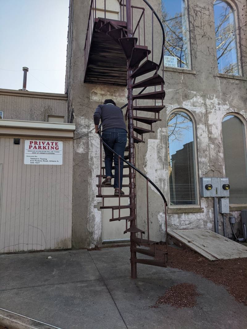 Image: The writer is climbing up a metal spiral staircase on the outside of a grayish walls. Photo by Andrea Black. 