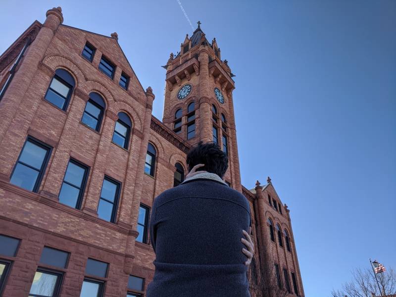 Image: The writer is facing backwards with hands wrapped around himself. He is standing in front of the Champaign County Courthouse, a red brick building with a clock tower. Photo by Andrea Black. 