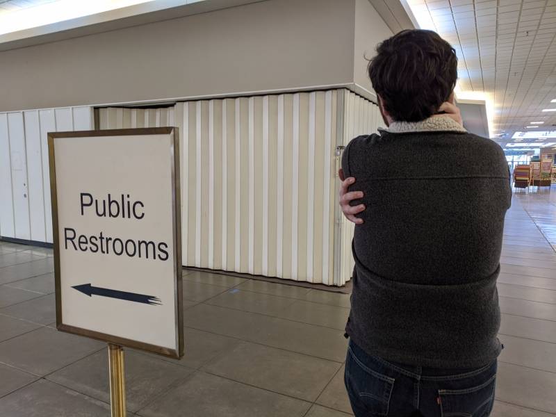 Image: The back of the writer with his hands wrapped around himself. He is standing in an empty mall with a public restrooms sign to his left. Photo by Andrea Black. 