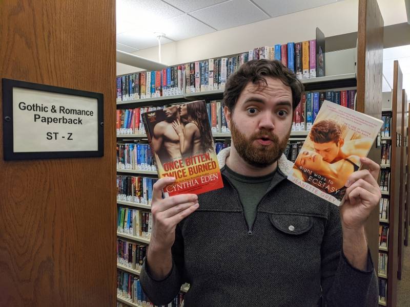 Image: The writer is standing between two library shelves, one of which is labeled Gothic & Romance Paperback. He is holding two books. Photo by Andrea Black. 