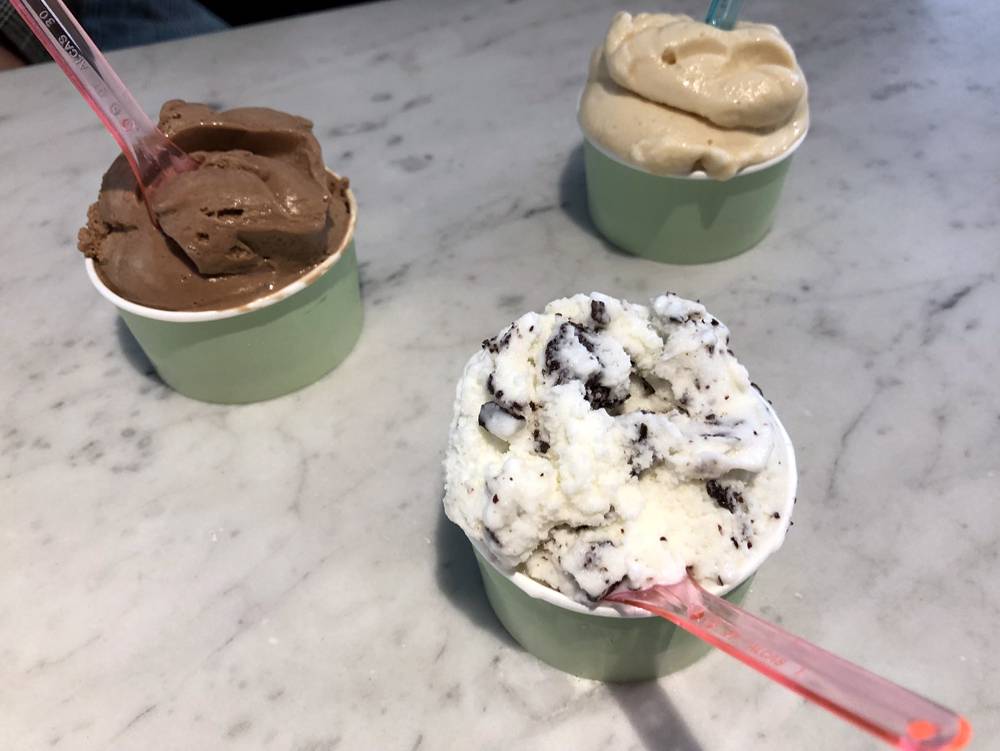 Three cups of ice cream in sage green paper cups sit on a marble countertop. Each cup has a plastic spoon stuck in it. One ice cream is chocolate, one is cookies and cream, and one is salted caramel. Photo by Jessica Hammie. 