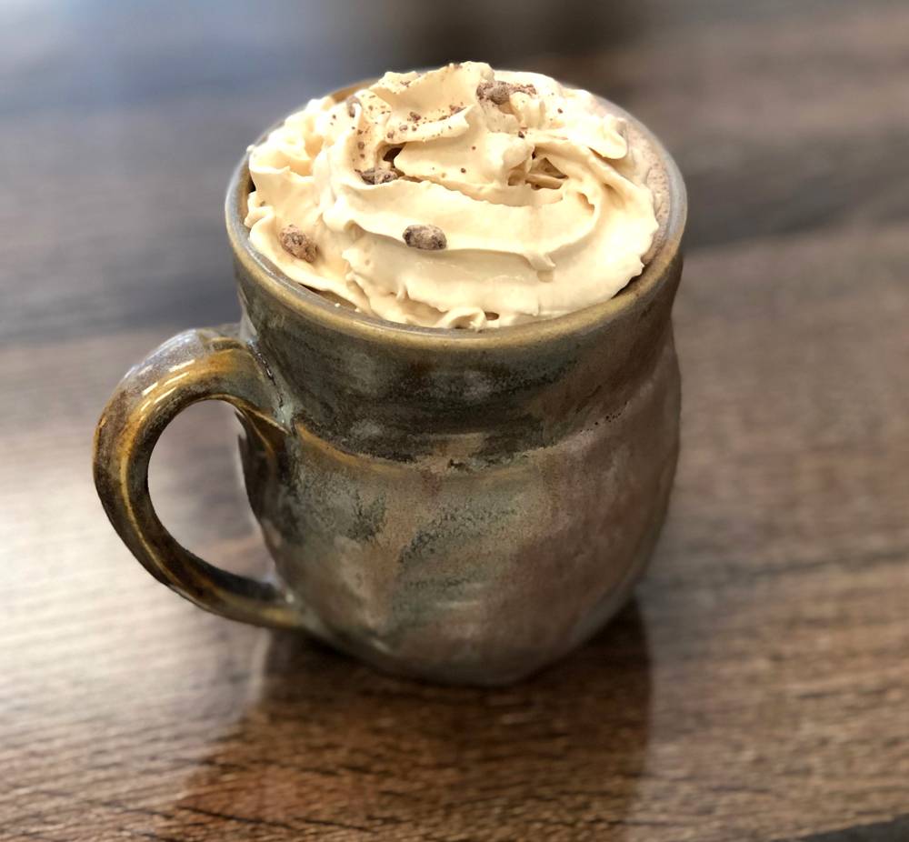 Image: A mug of hot chocolate at Bossa Nova Cafe sits on a gray wood table. The ceramic mug is gray-green, and a swirl of whipped cream sits above the rim of the mug. The whipped cream is garnished with small pieces of chocolate. Photo by Jessica Hammie.