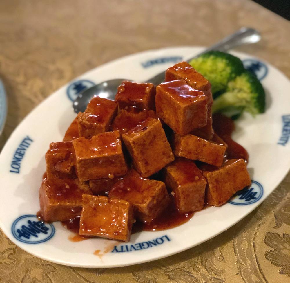 General Tso's tofu at Peking Garden. Large cubes of fried tofu are covered in a glossy, reddish brown sauce and served on a white plate with blue graphics. The plate is on a gold tablecloth. Photo by Jessica Hammie. 