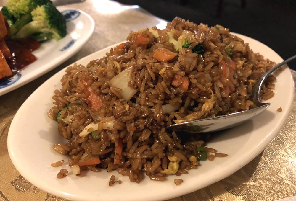 Image: Veggie fried rice at Peking Garden is served on an oval, white plate. A silver serving spoon is nestled in the side of the rice. The rice is brown and various small, chopped vegetables are mixed in. Photo by Jessica Hammie. 