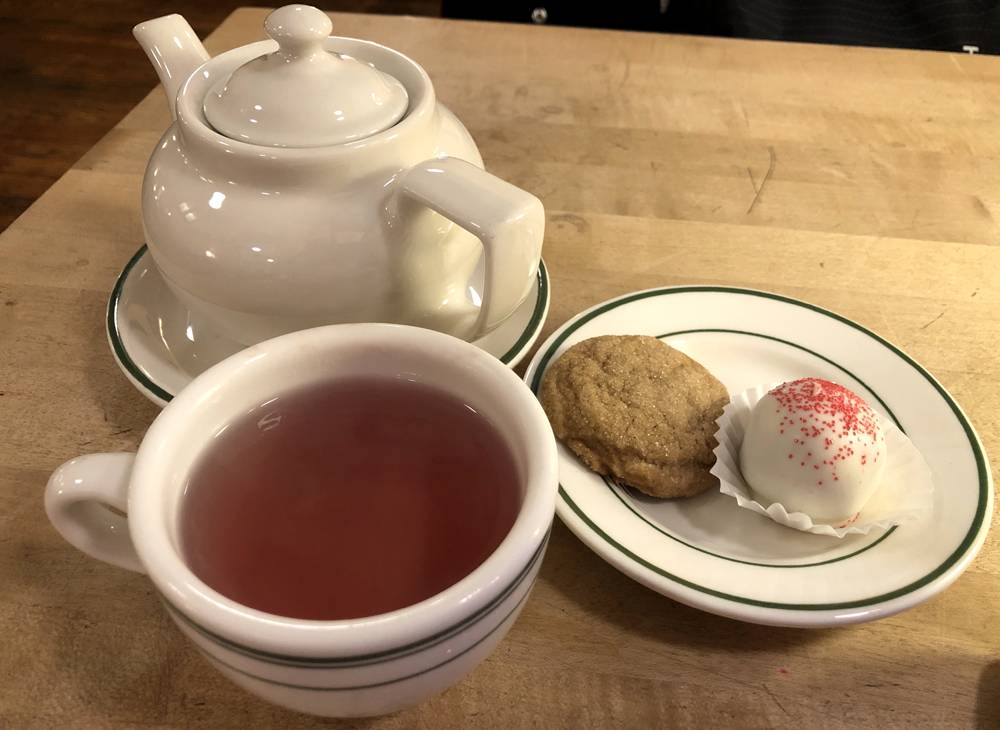 Image: A tea service at Cafe Kopi includes a white ceramic teapot on a white ceramic saucer with dark green trim on the edge, a white ceramic mug with dark green trim and a pinkish tea inside, and a white ceramic plate with dark green trim. On the plate are two cookies: one ginger snap, and one red velvet cake truffle dipped in white chocolate and dusted with red sprinkles. Photo by Jessica Hammie. 