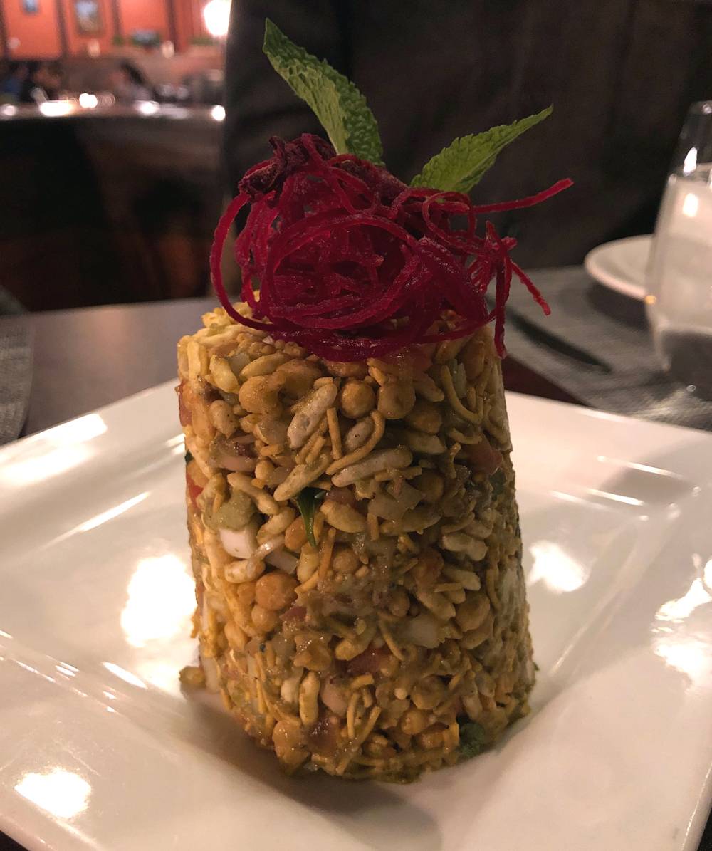 Avocado jhalmuri is stacked on a square white plate at Himalayan Chimney. The stack of avocado, puffed rice, and chickpeas is topped with very thin strands of raw beets and topped with a mint leaf. Photo by Jessica Hammie.