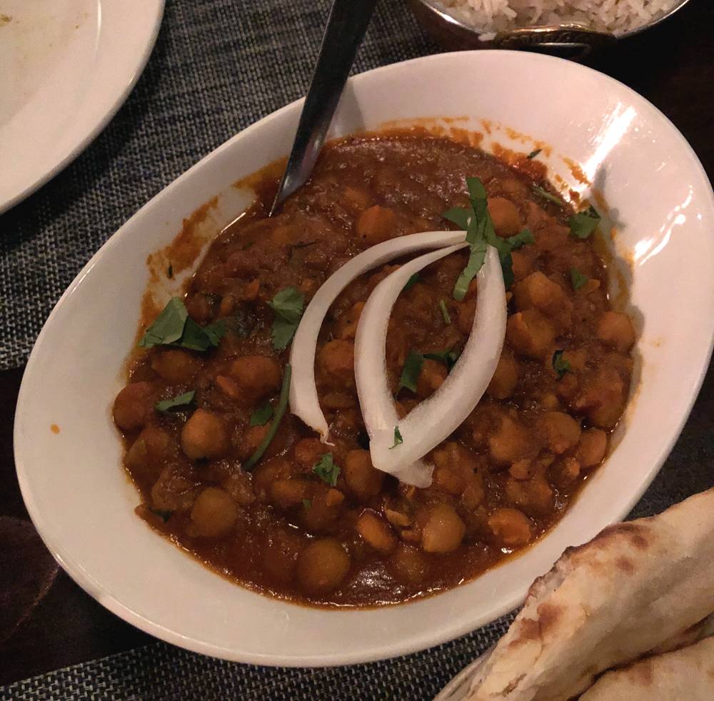 A bowl of chana pindi at Himalayan Chimney. Chickpeas in a reddish tomato sauce are garnished with three slivers of white onion an served in an oval white bowl. There is a metal spoon in the bowl. Photo by Jessica Hammie. 