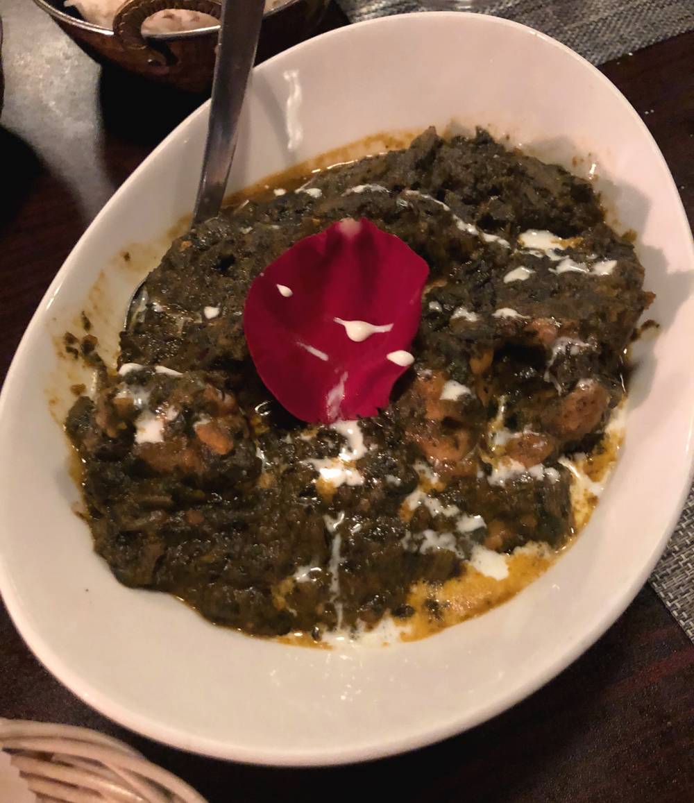 A bowl of chicken saag at Himalayan Chimney. Dark green spinach puree and pieces of chicken are garnished with a rose petal and drizzle of raita. It is served in an oval white bowl. photo by Jessica Hammie. 