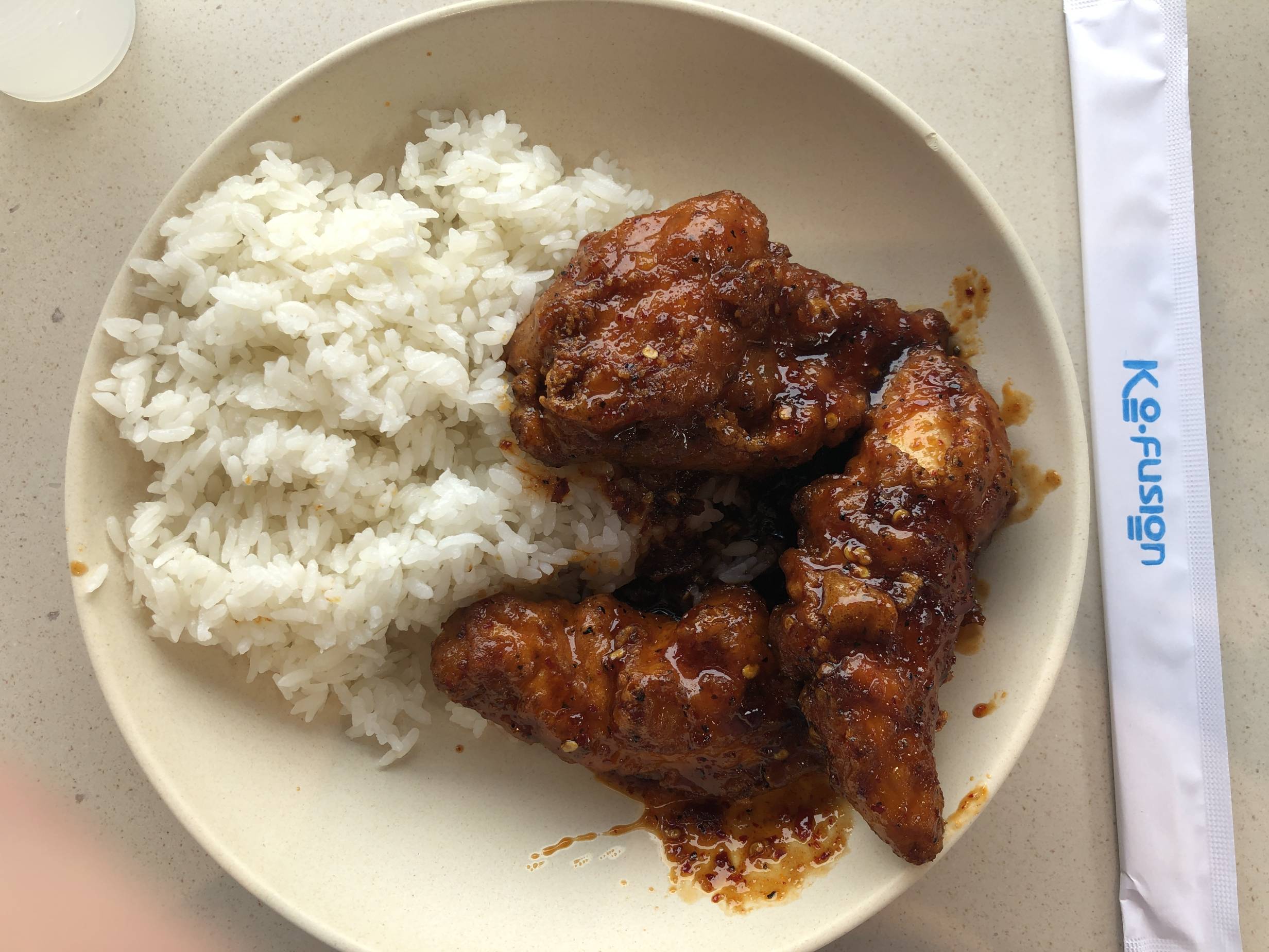 A plate of spicy fried chicken at KoFusion is served with white rice on a white plate. The chicken is a golden-red color with flecks of pepper flakes throughout the glossy sauce. There is a white paper-wrapped set of chopsticks to the right of the plate. Photo by Alyssa Buckley.