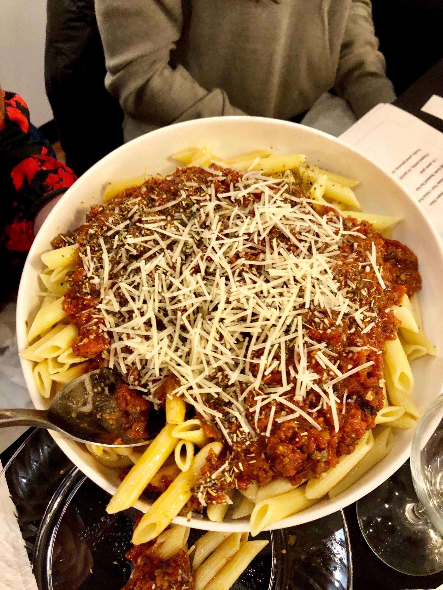 Image: A large, round, white bowl of pasta puttanesca with a silver serving spoon. The penne pasta is plain, and the red sauce sits on top. On top of the sauce are silvers of grated parmesan cheese. Photo by Rebecca Wells. 