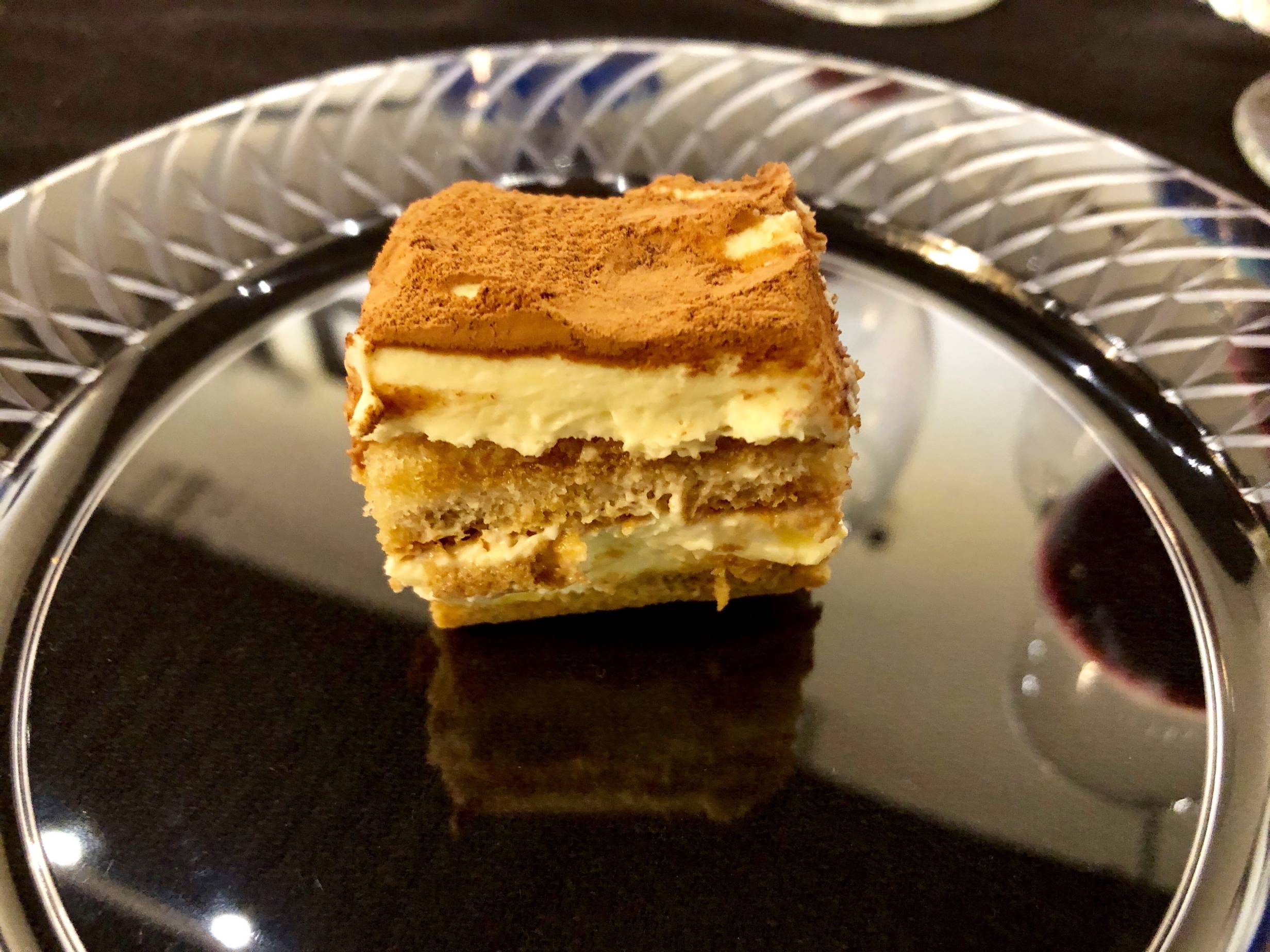 Image: A small slice of tiramisu sits on a round, clear plate on a black tablecloth-covered table. The tiramisu is layers of coffee-soaked lady fingers, and cream-colored mascarpone cheese. The top of the tiramisu is dusted with cocoa powder. Photo by Rebecca Wells. 