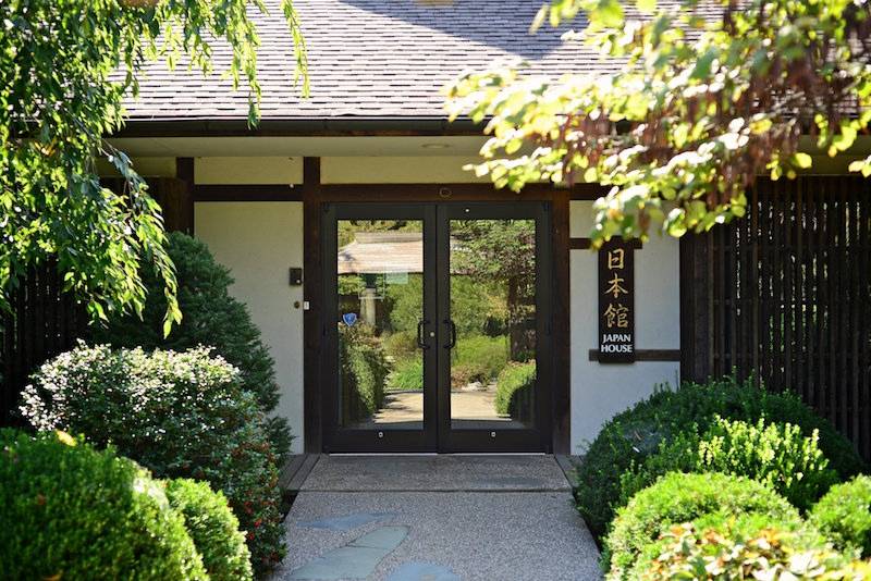 The entrance to Japan House. There are glass double doors, and the facade is white with brown wooden planks forming a grid around the entrance. There are green bushes and shrubs lining a walkway to the entrance. A brown rectangular sign to the right of the doors has Japanese characters in gold and says Japan House in white lettering. 