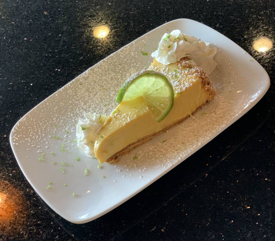 A piece of key lime pie at Sun Singer. A wedge of a yellow custard-based pie is served on a rectangular white plate. The pie is garnished with a slice of lime, whipped cream, and lime zest. The plate sits on a black table. Photo by Stephanie Wheatley. 
