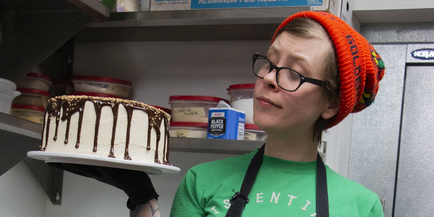 In a commerical kitchen, a person wearing a green t-shirt, black apron, black glasses, and an orange knit hat looks at a cake they hold to the side. The cake is cream colored with chocolate drips down the side. Photo by Steven Pratten.