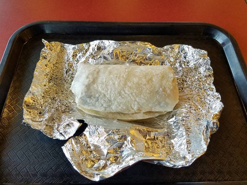 A Moe's burrito. Only the flour tortilla is visible. It sits on unwrapped tin foil on a dark, plastic cafeteria tray. Photo by Matthew Macomber. 