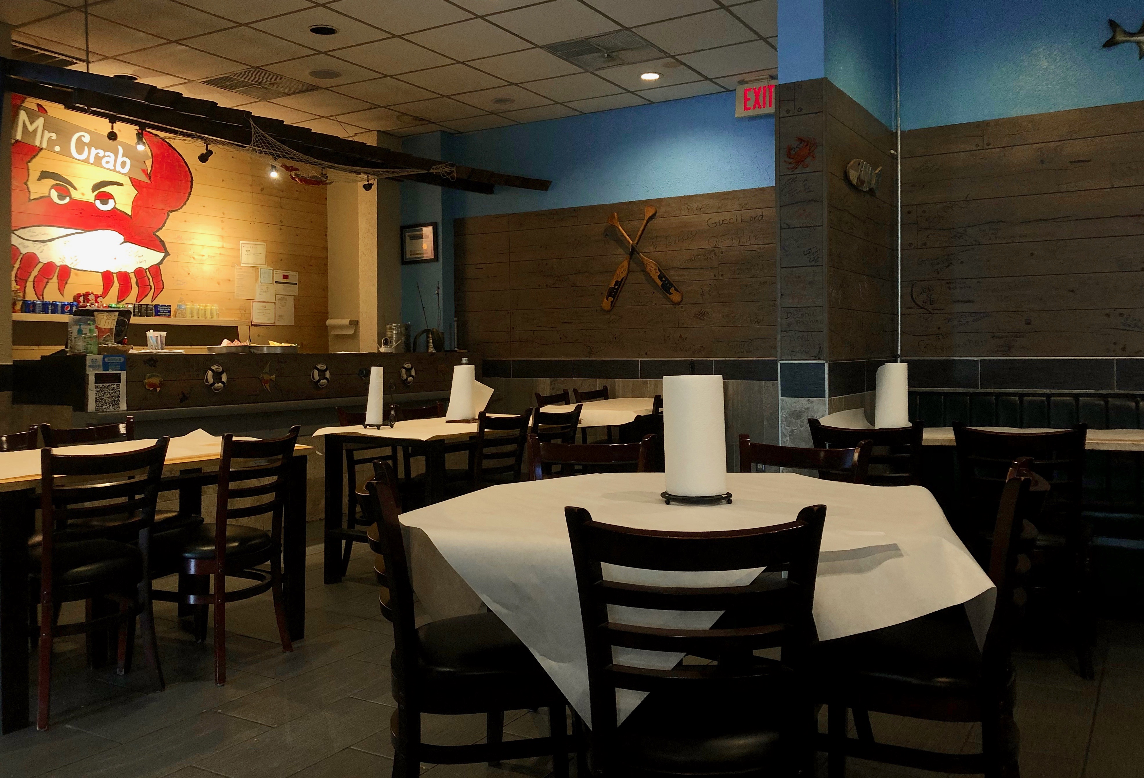 Image: The inside of Mr. Crab restauarant's dining room. Tables are lined with white paper. Paper towel rolls are on each of the tables. The top half of the walls are covered in shiplap. On the left side there is a mural of a crab with 