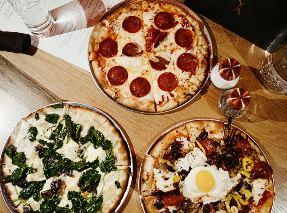 At NAYA, three pizzas are served on individual metal baking dishes. One pizza is pepperoni, one has white sauce and mushrooms, one is a supreme style pizza with an egg. The table is a light brown wood and there are salt and pepper shakers on the table. Photo by Anna Longworth. 