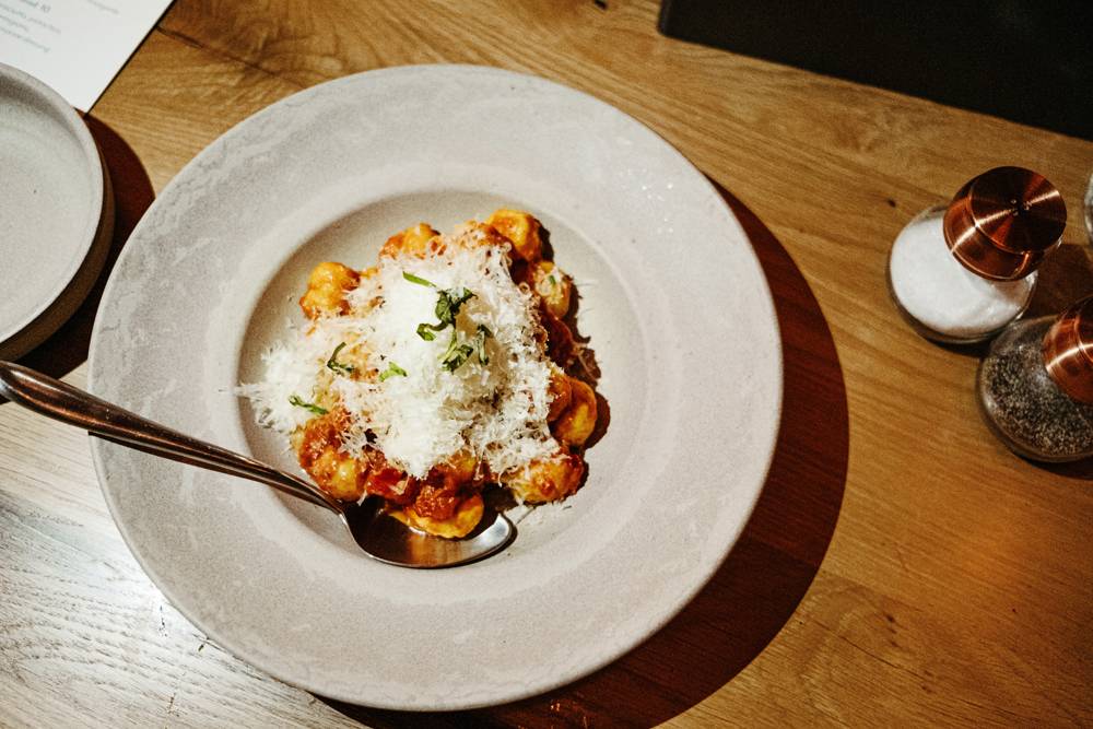 Daily gnocchi special at NAYA is served in a large, gray ceramic dish. The gnocchi is garnished very generously with grated parmesan cheese. A metal spoon sits in the dish. There are salt and pepper shakers on the light brown wood table. Photo by Anna Longworth. 