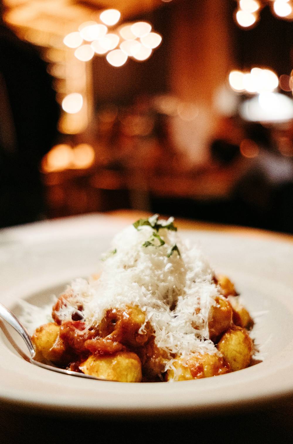 A side view of the daily gnocchi special at NAYA. Gnocchi are served in a large, gray ceramic dish. The gnocchi are garnished very generously with grated parmesan cheese. A metal spoon sits in the dish. Photo by Anna Longworth.