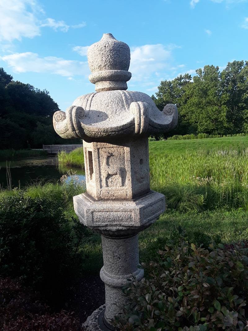 A carved concrete Japanese sculpture stands in a grassy area with a pond in the background.