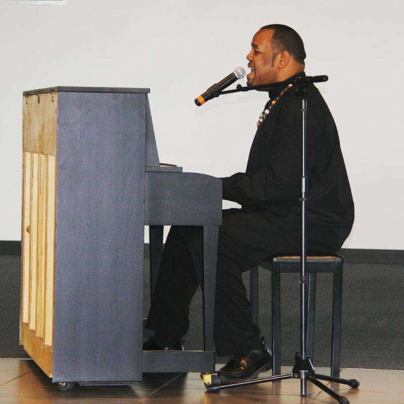 Image: A man is seated at a piano, playing and singing into a microphone. He is wearing a black shirt, black pants, and black shoes. Image from Parkland College Facebook page.