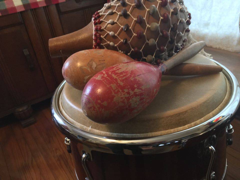 A wooden shaker with a beaded net around it sits on top of a drum along with a pair of maracas. Behind the drum, light shines through a window.