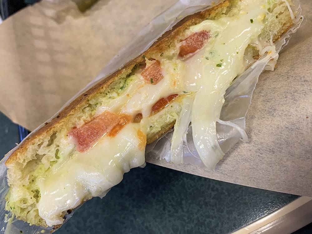 Image: The inside cross section of the pesto grilled cheese at Baldarotta's. Thin and crispy bread contains oozing white cheese, green pesto, and diced tomatoes. The half of the sandwich is wrapped in white wax paper and sitting on brown paper. Photo by Zoe Valentine. 