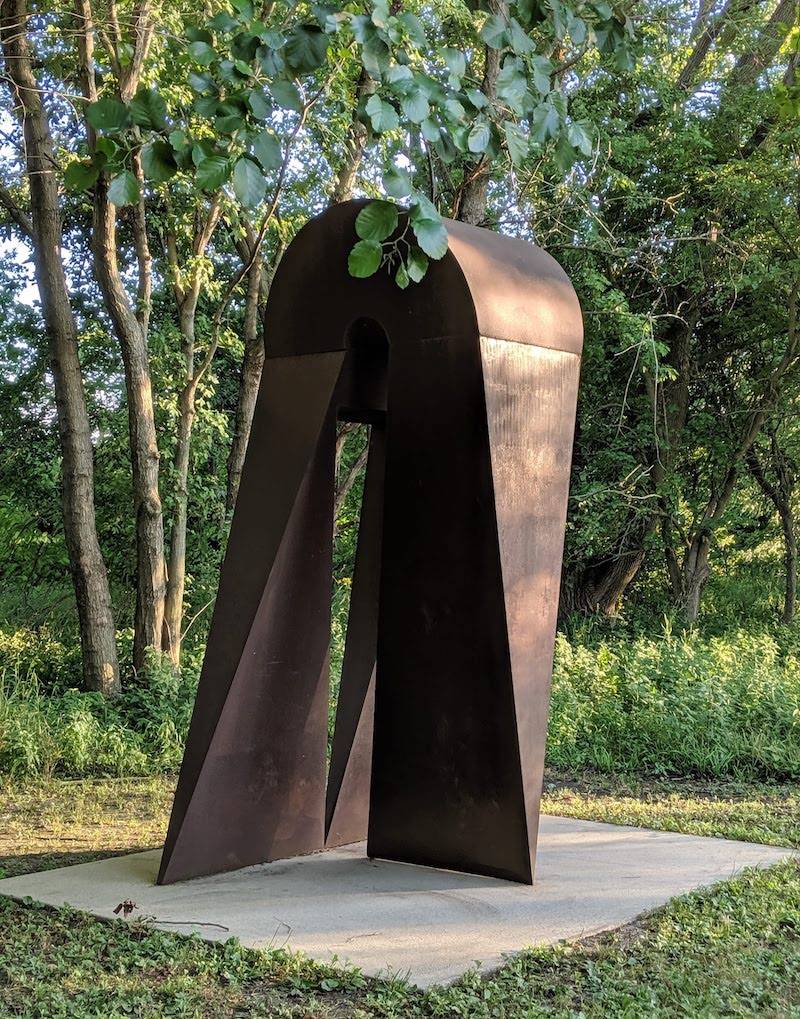 A metal sculpture that is rounded at the top and has three legs that come down, getting narrower as they get closer to the ground, coming to a wedge shape at the bottom. It sits on a slab of concrete and is surrounded by green grass and trees. Photo by Katriena Knights.