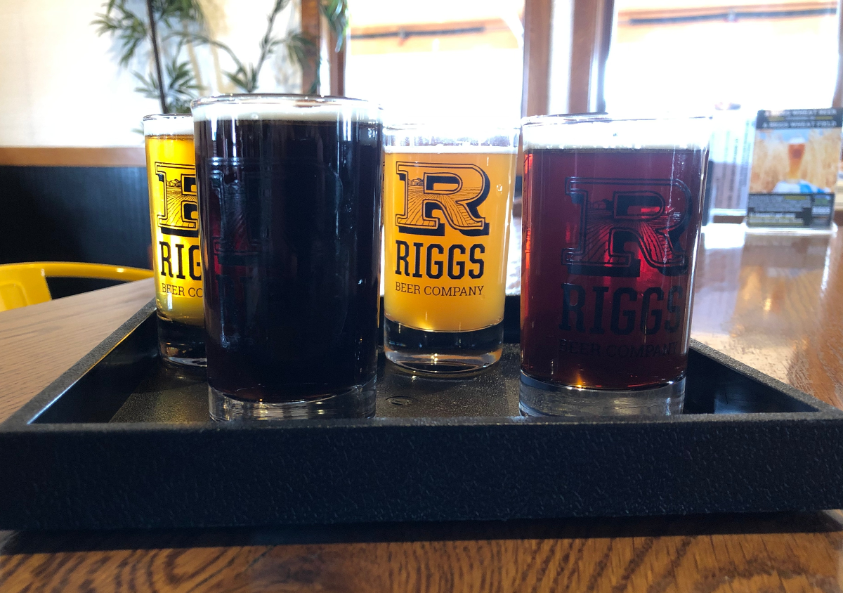 Four beer glasses sit in a black rectangle tray on a wooden bar at Riggs. Three of the glasses show 