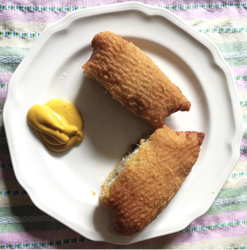 A square potato knish is cut in half and sits on a round white plate. There is a squirt of yellow mustard on the plate. The plate sits on a pastel-colored striped tablecloth. Photo by Jessica Hammie. 