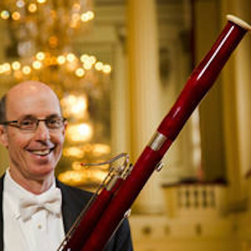 Image: Henry Skolnick smiles in a tuxedo and thin-framed glasses while holding his bassoon at a diaganol to his body. Behind him, a grand hall with pillars and chandeliers out of focus. Image from Skolnick's educator's profile with Interlochen music camp.