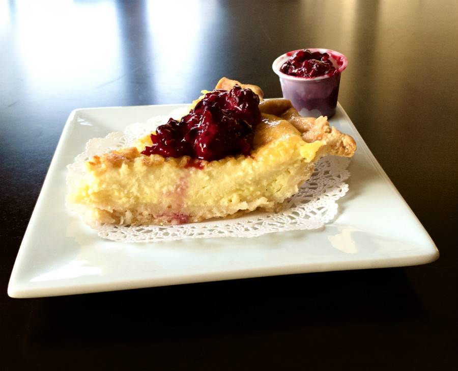 Buttermilk pie with berries on top at Lucky Moon Pies. A slice of yellow pie sits on a white paper doilie on a square, white plate. There is a small plastic condiment bowl with extra berries on the side. Photo by Stephanie Wheatley. 