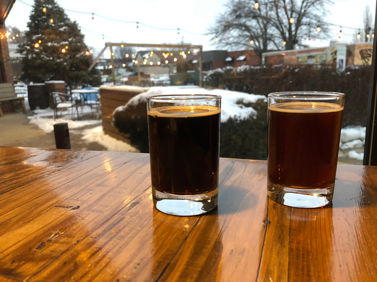 Two glasses of dark brown beer sit on a wooden table. In the background are string lights and a dusting of snow on trees and bushes at Tripych's outdoor area. Photo by Jesus Barajas.