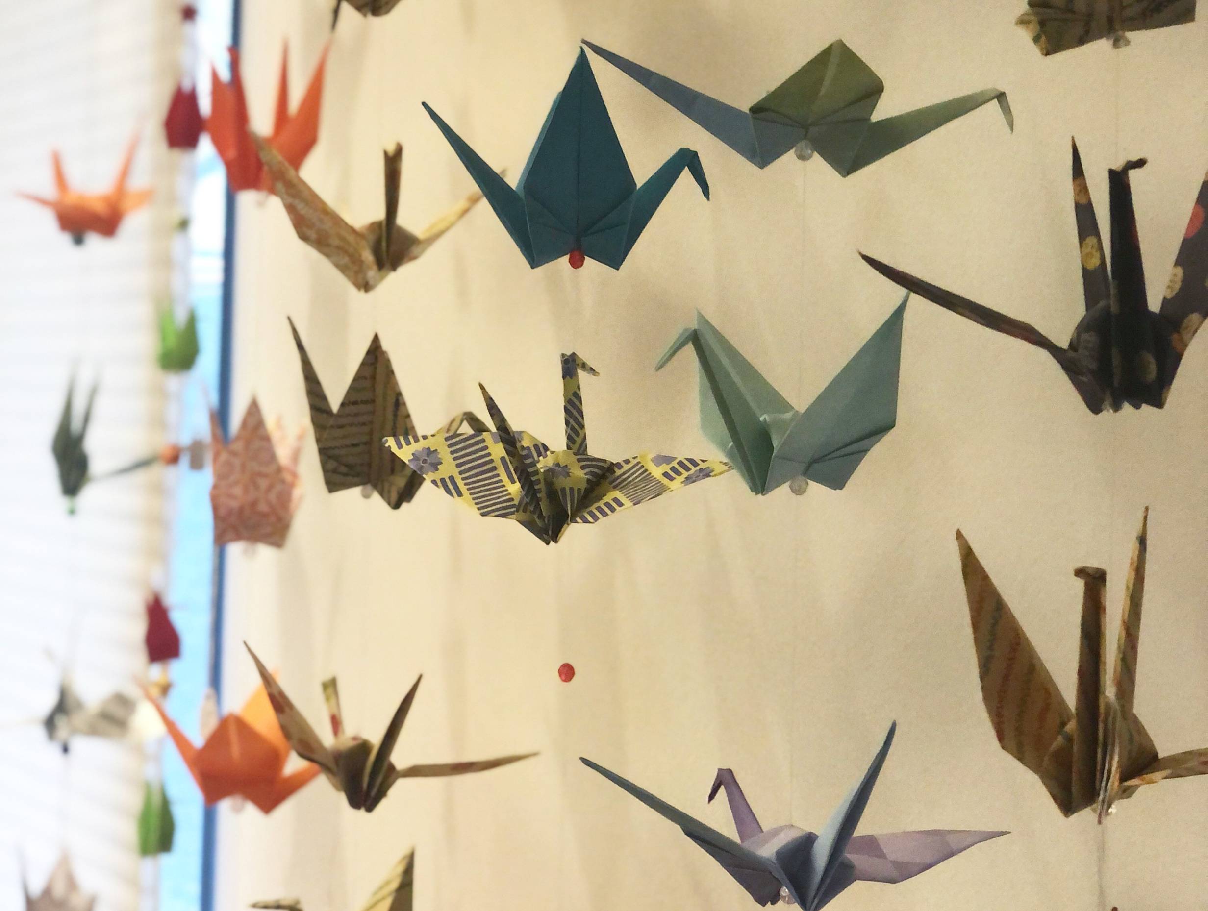 Paper cranes in various colors hang in vertical lines on string along a white wall at Japan House. Photo by Alyssa Buckley.