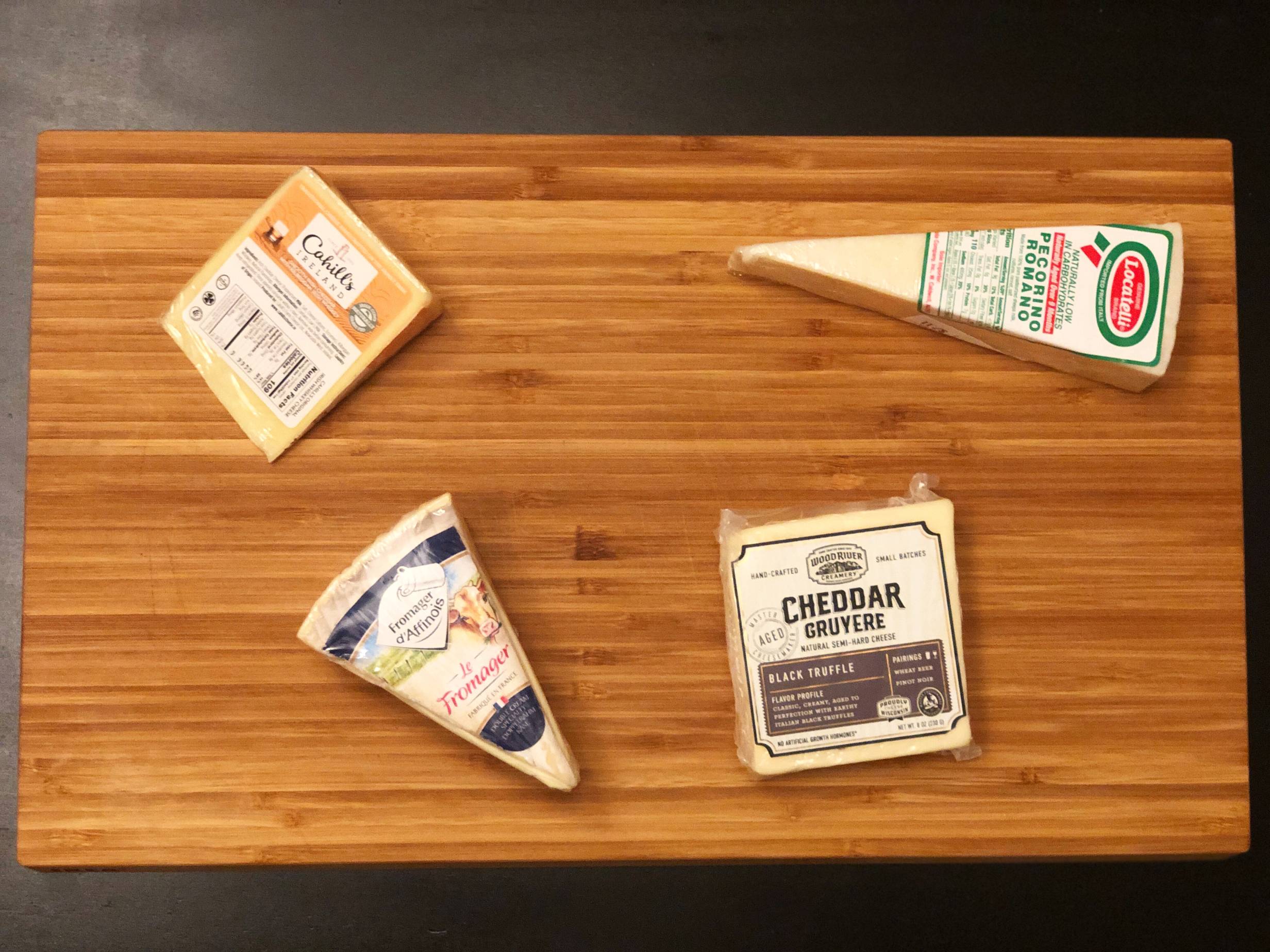 A wooden cutting board with four cheeses. The top left is a square of Cahill, top right is a skinny triangle of Pecorino. Bottom left is a wedge of Fromager dâ€™Affinois, and the bottom left is a square of Cheddar. All are in the plastic packaging with labels in view. Photo by Alyssa Buckley.