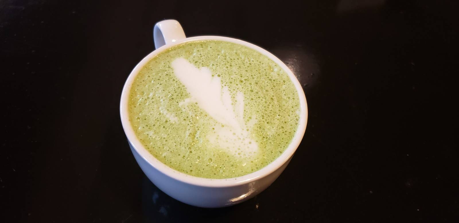 A matcha latte at Cafeteria & Company. Green matcha latte features a white feather design in the milk foam. The mug is white and sits on a black table. Photo by Elaine Sine. 