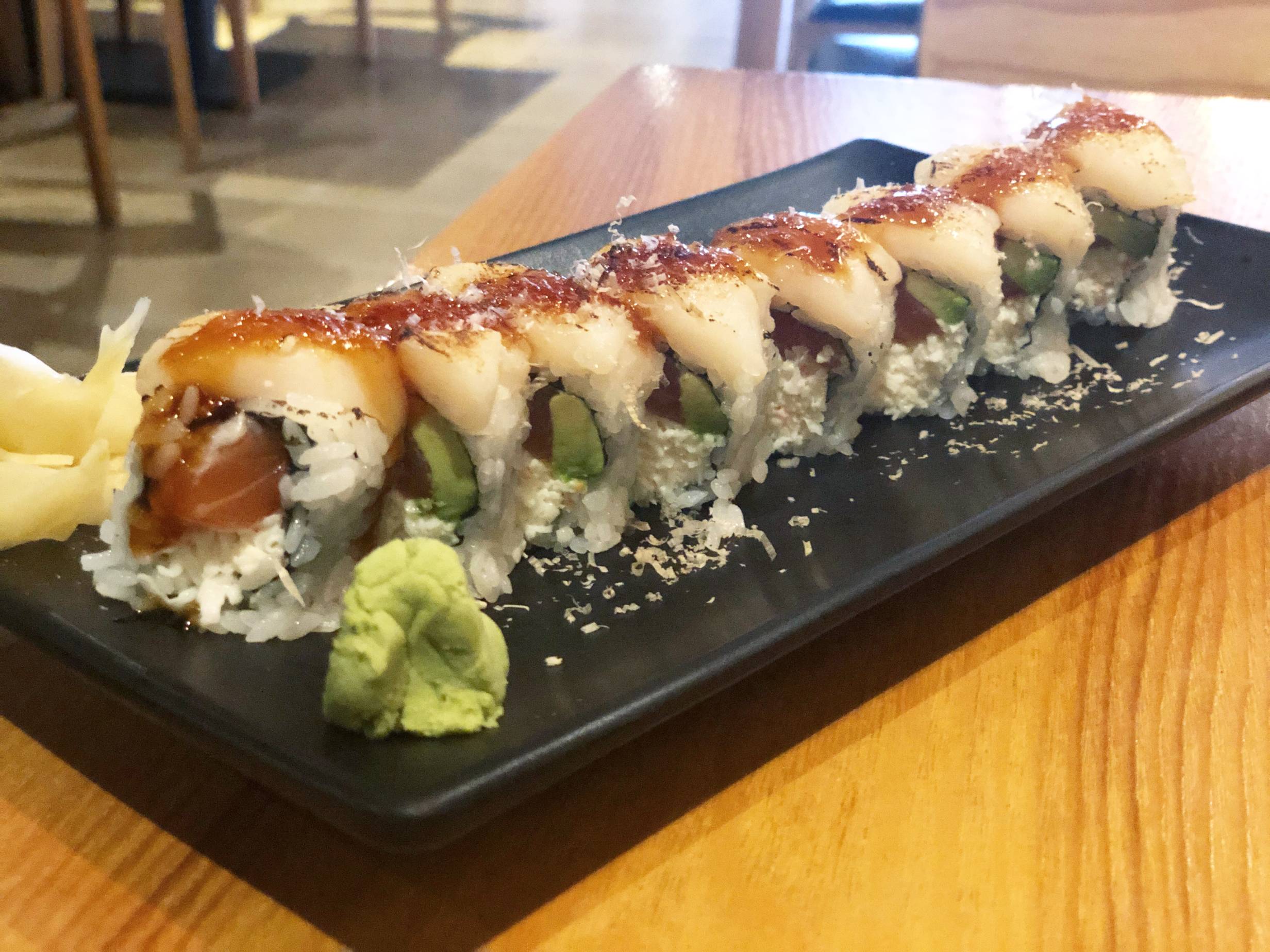 An urarmaki roll sit on a black rectangular plate on a wooden table. The sushi roll has white sushi rice wrapped around nori with raw salmon, avocado, and crab inside. On top of the roll, there is a small piece of scallop with a dark sauce and a dusting of fish flakes. Next to the roll on the left side of the plate is a pile of yellow pickled ginger and a small mound of green wasabi. Photo by Alyssa Buckley.