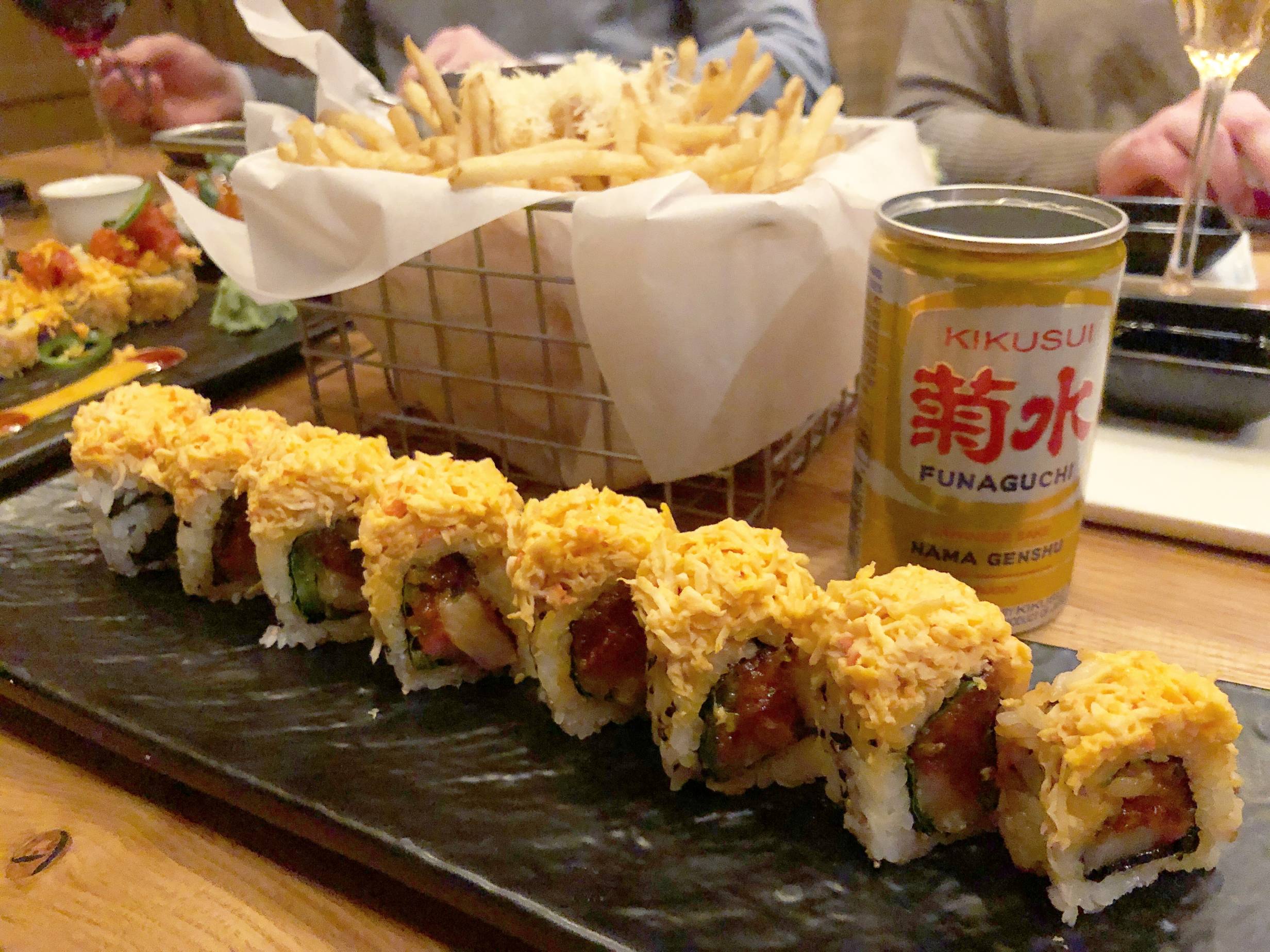A tuna and yellowtail sushi roll topped with spicy crab sits on a black, rectangular plate. Behind it, there is a metal basket lined with parchment paper and fries topped with Parmesan sticking out of the basket. Beside the fries is a small, gold can of Funaguchi sake on a wooden tabletop. In the background of the image are other diners at Miga. Photo by Alyssa Buckley.
