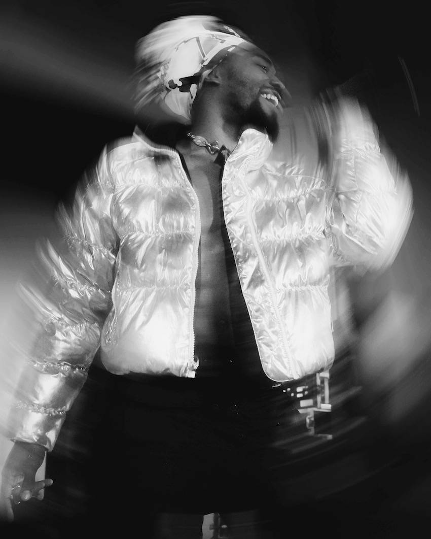 In this black and white photo, a member of EarthGang raps into a microphone. He wears dark pants and a bright, puffy coat that is unzipped to reveal a bare torso. His movement is captured in the long exposure of the photo.